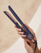A navy blue and rose gold flat iron held in a person's hand against a soft beige background.