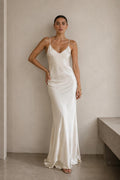 A woman modeling a floor-length white satin slip dress with thin spaghetti straps and a V-neckline, standing in a minimalist room with a textured beige wall and concrete floor.