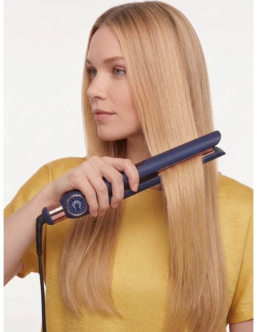 A woman with long blonde hair uses a navy blue and rose gold flat iron on a section of her hair, wearing a yellow top against a plain white background.