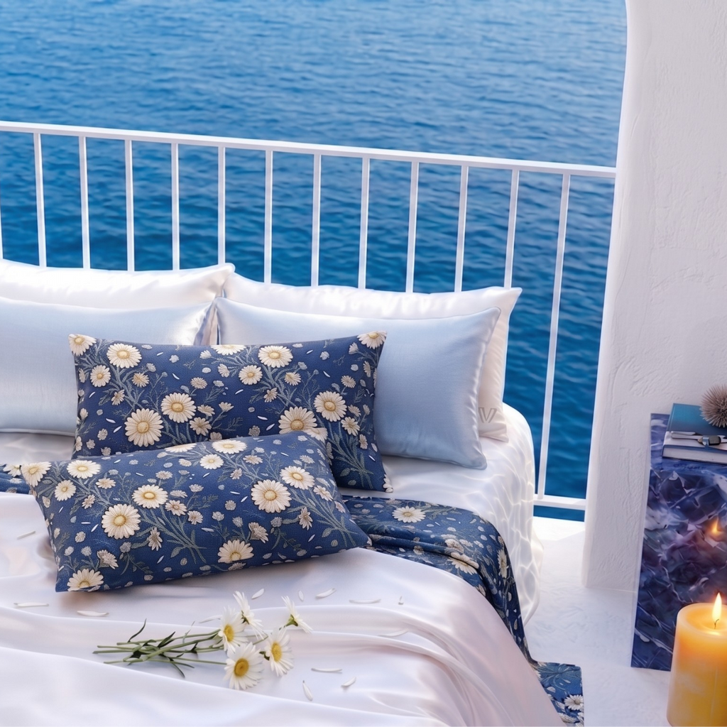 A styled bed with white bedding and navy blue floral pillows and duvet, set on a balcony overlooking a blue ocean. Decorative blue marble side tables, a lit candle, and small plants are placed around the bed.