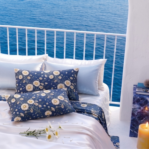 A styled bed with white bedding and navy blue floral pillows and duvet, set on a balcony overlooking a blue ocean. Decorative blue marble side tables, a lit candle, and small plants are placed around the bed.
