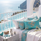 A styled daybed with white and turquoise floral pillows and a matching throw blanket, set on a balcony overlooking a bright blue ocean and coastal cliffs.